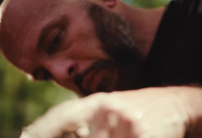 A male artisan with tattoos concentrates intensely as he molds a piece of clay on a spinning pottery wheel, with a rack focus from his hands to his face