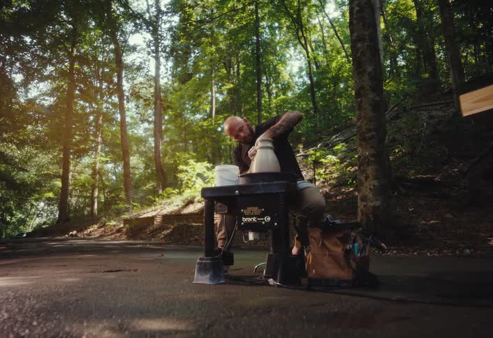 A bald, tattooed man skillfully shapes clay on a pottery wheel in a serene, sun-dappled forest
