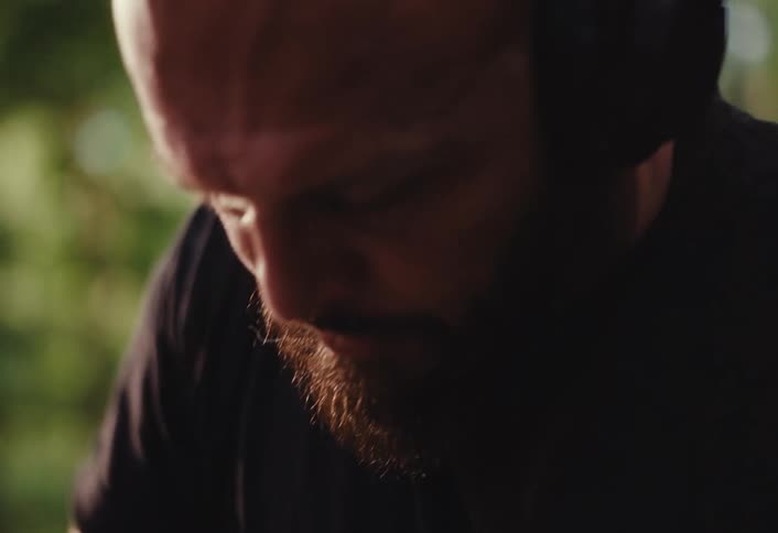 A tattooed artisan with a beard and headphones concentrates on shaping wet clay on a spinning potter's wheel