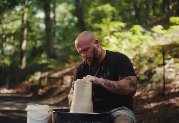A bearded and tattooed potter carefully shapes a clay pot on a pottery wheel in a tranquil forest setting