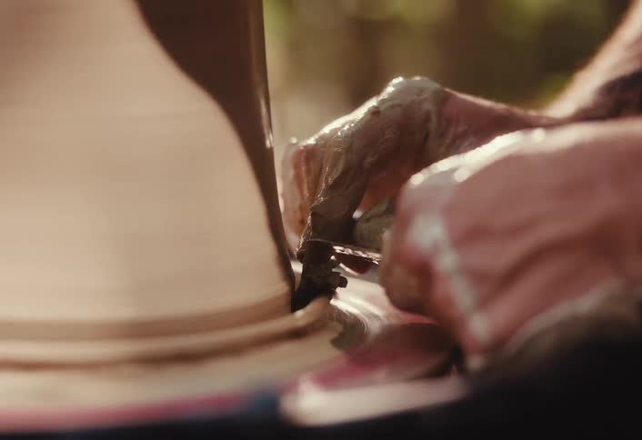 Slow motion close up of a potter's hands using a tool to shape the base of a spinning clay vessel on a pottery wheel