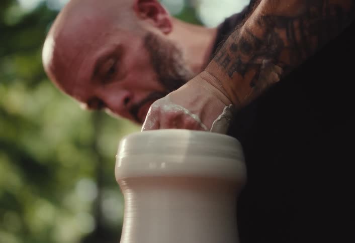 A bald, tattooed male artisan with a beard intensely focuses on shaping a piece of wet clay on a spinning pottery wheel in his outdoor workshop