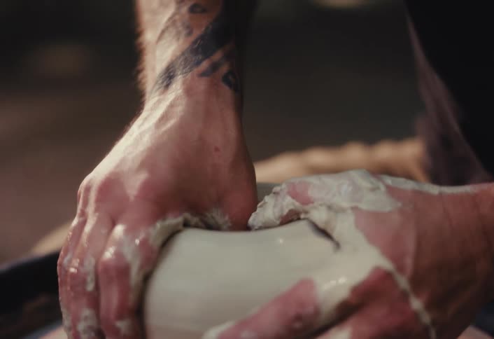 A tattooed artist with a beard intensely focuses while shaping a piece of wet clay on a spinning pottery wheel
