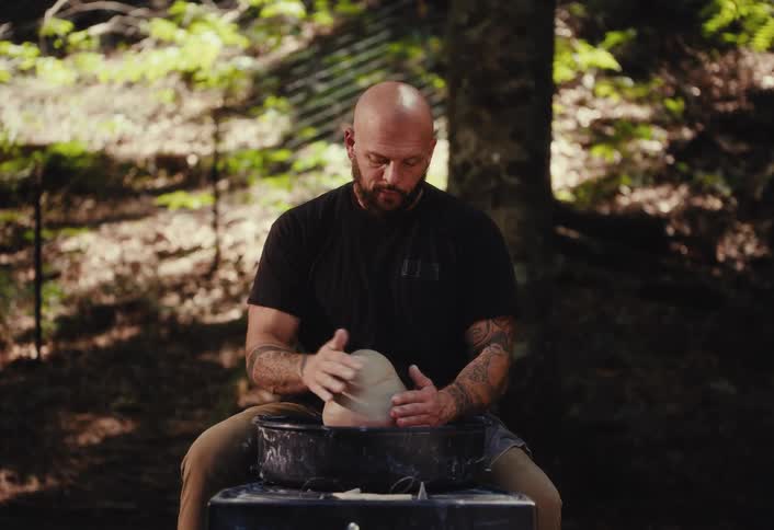 A bald, tattooed potter works on a lump of clay in the forest, then stops and gazes seriously at the camera