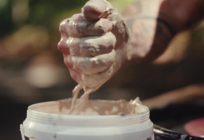 Slow-motion close-up of a tattooed hand dipping into a tub of wet clay and squeezing it into a dripping fist