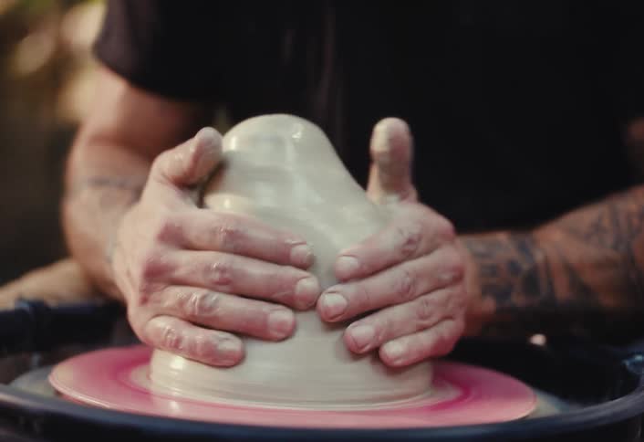 A bearded man with headphones and tattoos focuses on shaping a lump of clay on a spinning pottery wheel outdoors