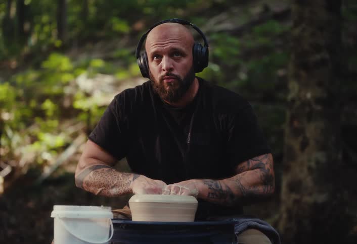A focused tattooed potter with headphones works with clay on a pottery wheel in a serene outdoor forest setting