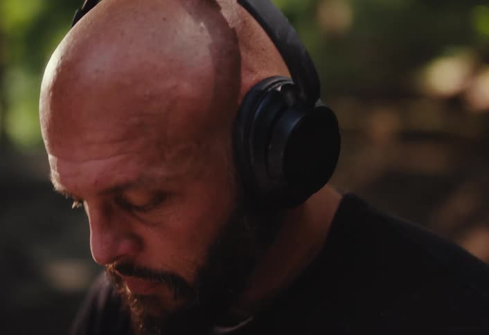 A close-up slow-motion shot shows a bald man with a beard putting on headphones in a park