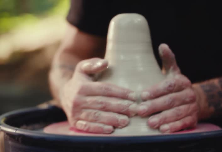 A bearded, tattooed man wearing headphones intently shapes a piece of clay on a potter's wheel in an outdoor setting