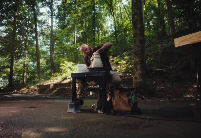 An artist with a beard and tattoos crafts pottery on a wheel in a peaceful, sunlit forest