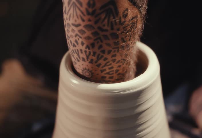 A tattooed potter skillfully shapes the inside of a spinning clay vessel with a wet sponge