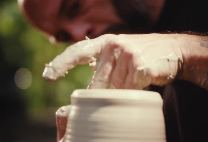 A focused, bearded potter skillfully shapes a turning piece of wet clay on a pottery wheel in slow motion
