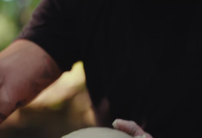 A focused, bearded potter wearing headphones skillfully shapes a piece of wet clay on a spinning wheel in an outdoor setting