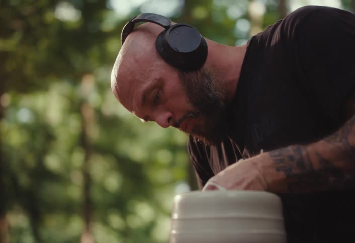 A tattooed artist with headphones concentrates on shaping clay on a pottery wheel in a peaceful forest setting