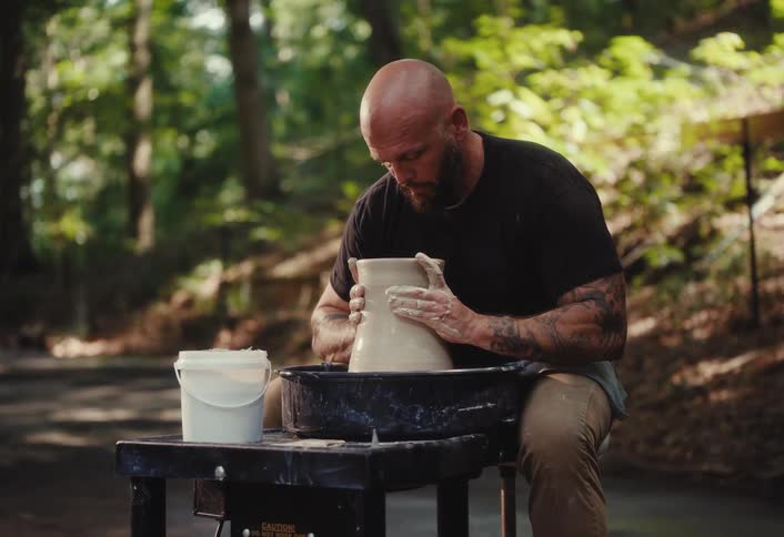 A focused, bald man with a beard and tattoos skillfully molds a piece of clay into a pot on a pottery wheel outside