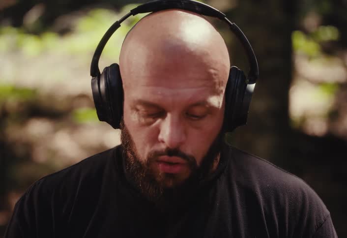 A bald, bearded man with tattoos meditates with headphones in the forest, then opens his eyes with an intense stare