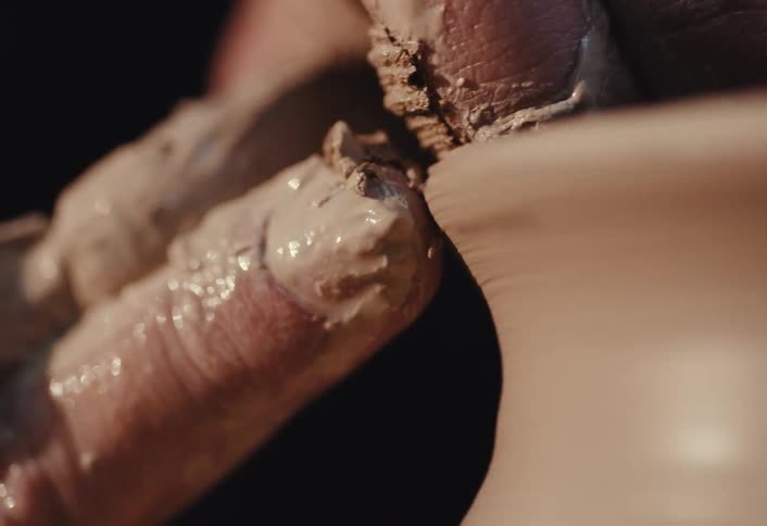 A macro shot shows a potter's hands covered in clay meticulously shaping the rim of a spinning vessel on a potter's wheel