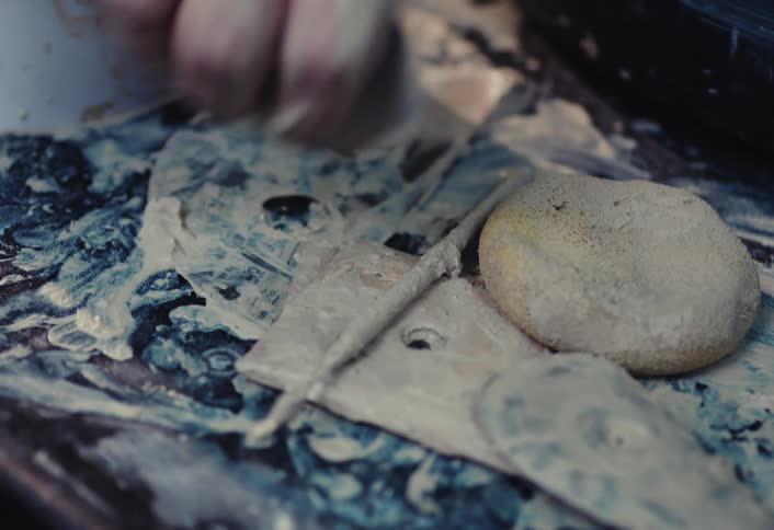 A close-up of a potter's messy workstation, with a hand reaching for a clay-covered tool among other instruments and a sponge