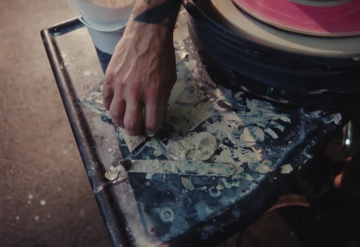 A potter's hand sorts through wet clay scraps and tools on a messy workspace next to a pottery wheel