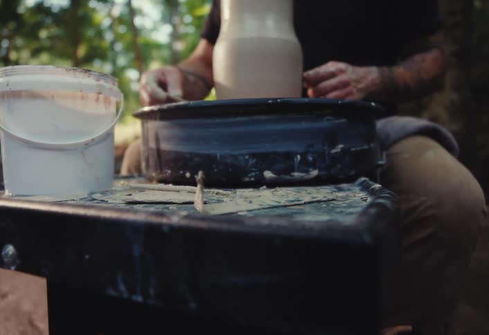 A potter with tattooed arms cleans up excess clay while working on a pottery wheel in a serene outdoor setting