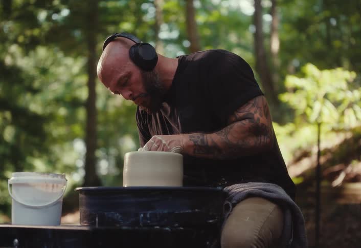 A focused, tattooed man wearing headphones carefully shapes clay on a potter's wheel in a tranquil outdoor forest setting