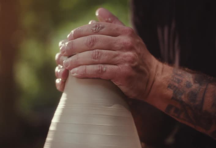 A close-up of a potter's tattooed hands skillfully shaping wet clay on a spinning pottery wheel