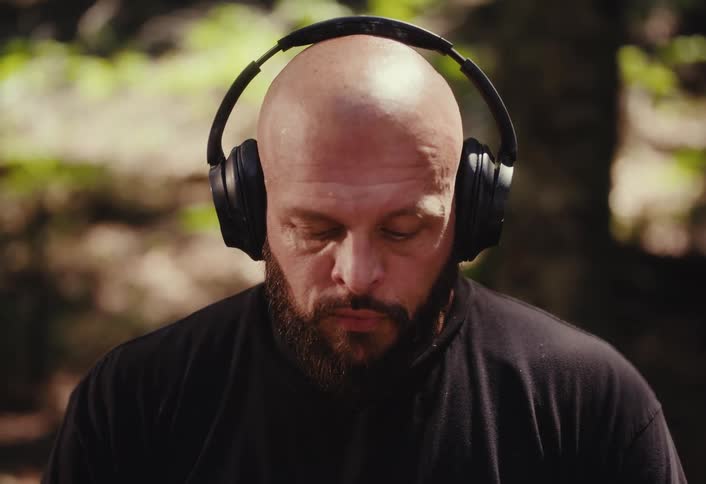 A bald, bearded man with tattoos meditates in a sunlit forest, putting on headphones before opening his eyes with a sudden, intense gaze