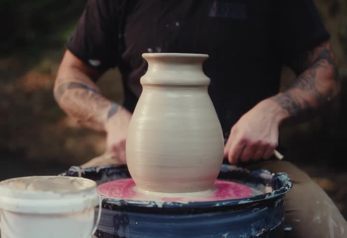 A close-up of a tattooed potter's hands skillfully using a wire to separate a finished clay vase from the pottery wheel