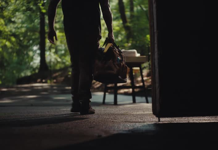A tattooed craftsman walks out of the shadows into a sunlit forest clearing to begin his work at a workbench