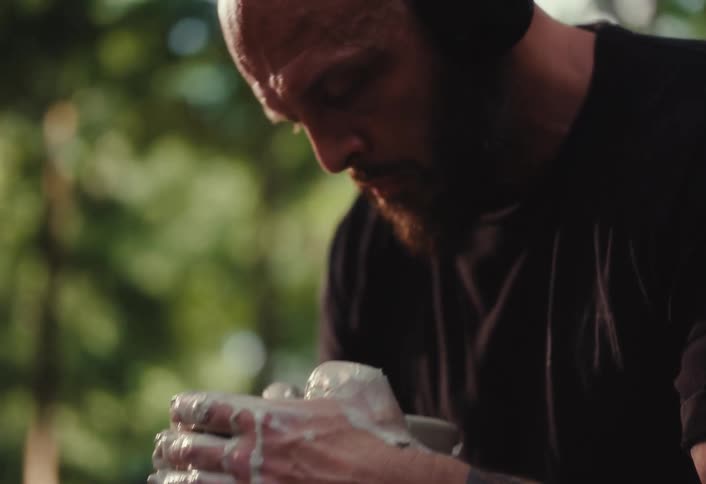 A focused bald man with a beard and headphones meticulously sculpts clay on a pottery wheel in a tranquil outdoor setting