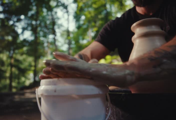 Close-up of a tattooed potter's hands cleaning a sponge over a bucket in a sunlit forest