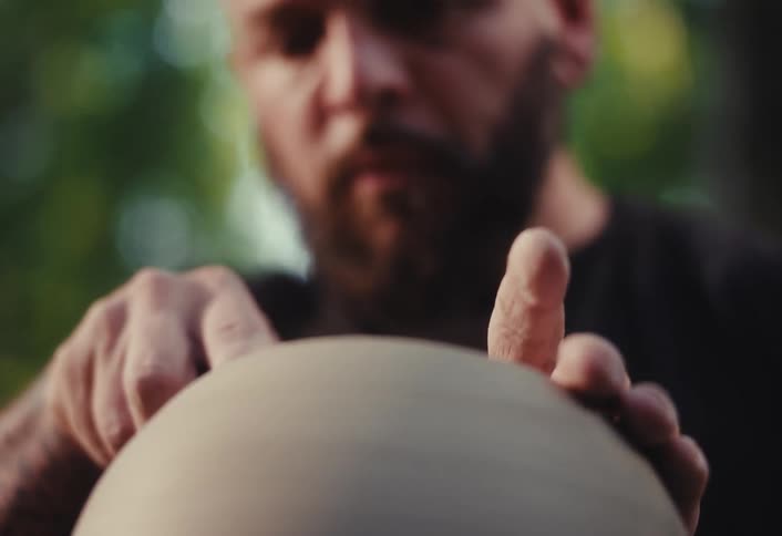 A skilled male potter with a beard meticulously shapes a piece of wet clay on a spinning pottery wheel in a close-up shot