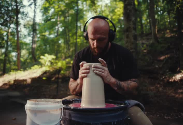 A tattooed man wearing headphones intently shapes a piece of clay on a pottery wheel in a peaceful outdoor forest setting