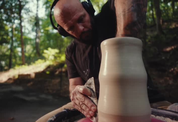 A potter with headphones carefully shapes a clay pot on a wheel in a peaceful outdoor wooded area