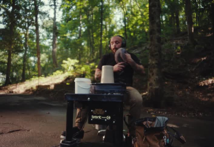 A tattooed artist with headphones wipes his face before beginning to shape clay on his pottery wheel in a serene forest setting