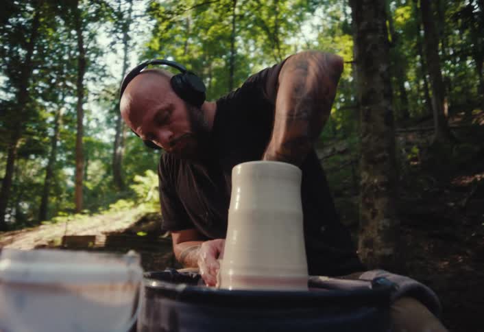 A focused, tattooed man wearing headphones shapes a piece of clay on a pottery wheel in a tranquil forest setting