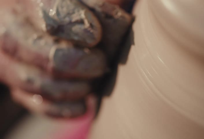 Extreme close-up of a potter's hands covered in wet clay shaping a pot on a spinning wheel