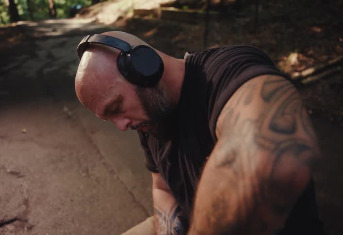 A focused, tattooed man wearing headphones skillfully shapes a clay pot on a spinning pottery wheel outdoors