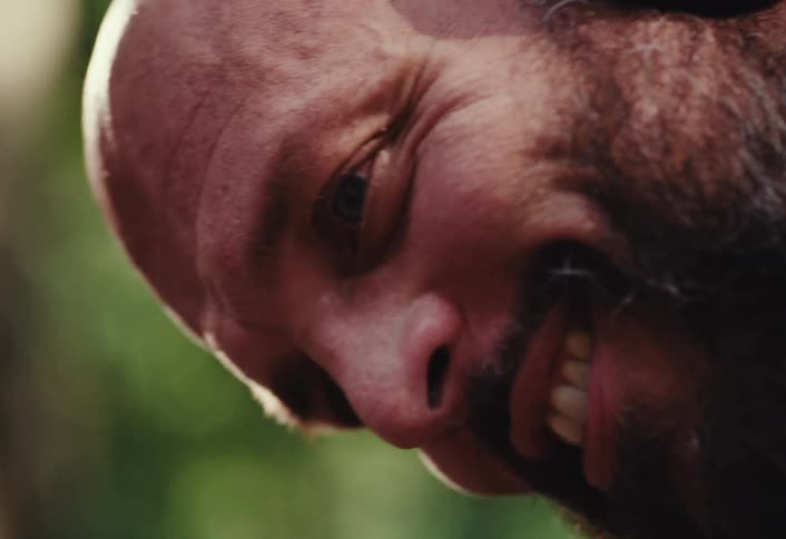 An intense vertical close-up of a bald, bearded man's face as he slowly transitions into a wide, menacing grin