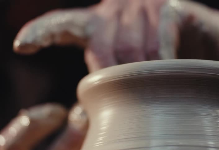 A potter carefully shapes the top edge of a spinning clay pot with their hands in slow motion