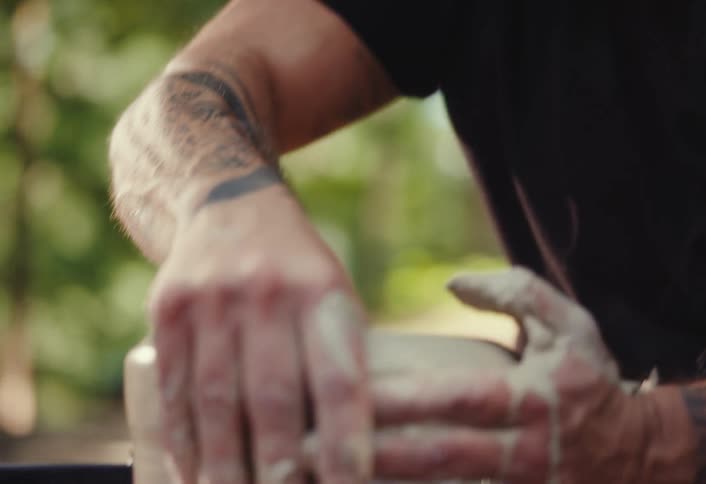 A focused, bald man with a beard and headphones works with his tattooed hands to shape clay on a spinning potter's wheel outdoors