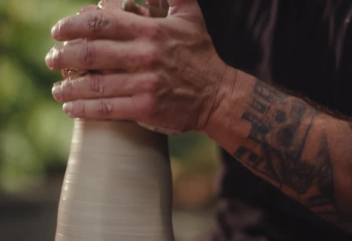 Close-up shot of a focused, bearded man with tattoos shaping wet clay on a spinning pottery wheel in a natural outdoor setting