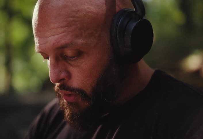 A bald, bearded man with tattoos puts on headphones in slow motion with a focused expression