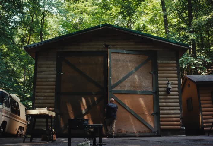 A bearded man works on a project outside his rustic log barn, closing the large doors before returning to his workbench in the woods