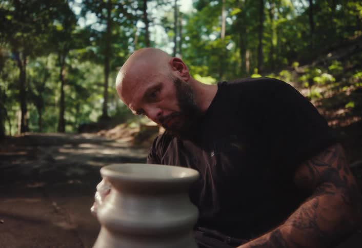An artisan potter carefully molds a spinning clay vase with his hands and a sponge on a pottery wheel in a tranquil forest setting