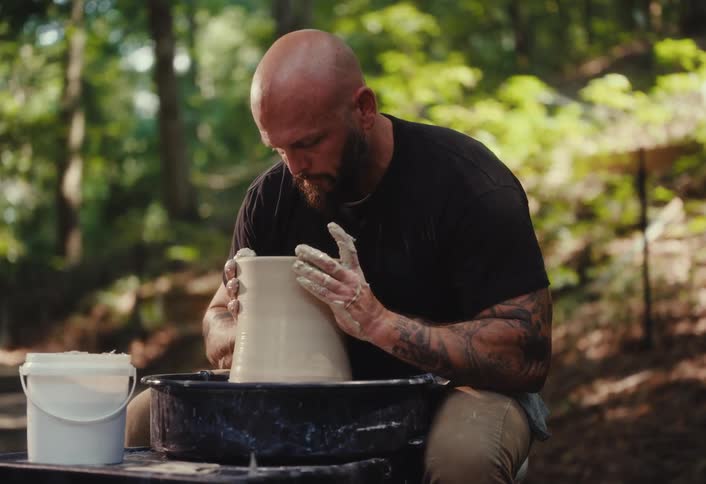 A bearded, tattooed man sits in a forest and carefully shapes a piece of pottery on a spinning wheel