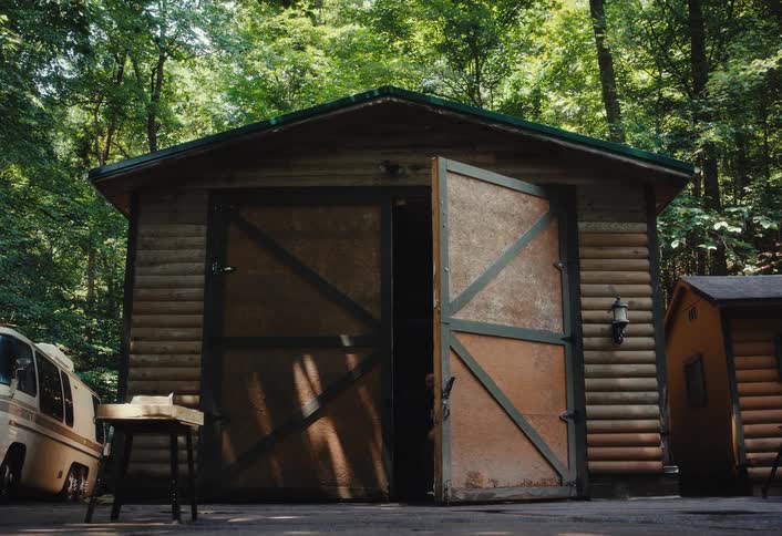 A man walks into frame, opens a rustic wooden shed in the woods, retrieves a chainsaw, and walks back into the shed