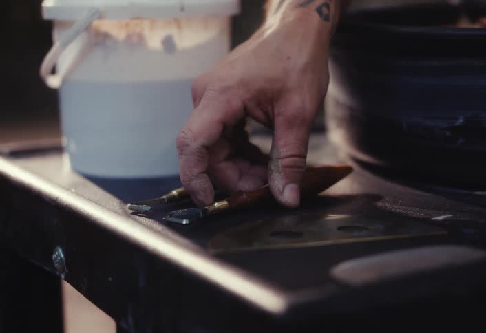 A close-up of an artist's hand, covered in clay, picking up a wooden-handled sculpting tool from a workbench