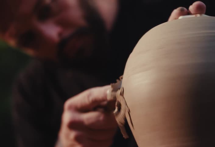Close-up slow motion shot of a craftsman's hands using a metal tool to shave and shape a spinning clay pot