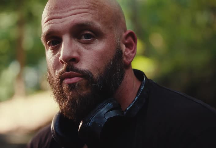 Close-up portrait of a serious bald man with a beard and headphones, looking intensely into the camera in a natural outdoor setting